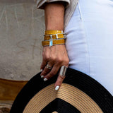 Yellow leather wrap bracelet with bold buckle, stacked with metal bangle, worn on a hand holding a black and tan hat.