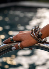 Unique hammered bangle bracelet in silver stacked with other bangles, worn on a hand resting on a railing by a pool.