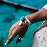 White leather wrap bracelet highlighting a chunky turquoise resin stone, paired with a silver half cuff, worn on a wrist.