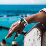 White leather wrap bracelet featuring a turquoise resin stone and a chunky metal cuff, worn on a hand by the water.