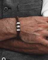 A close-up of a man's forearm wearing a dark brown leather bracelet with four silver-toned rectangular inlay beads, styled over a gray textured vest.