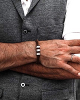A close-up of a man's crossed arms showing a brown leather bracelet with three silver-toned rectangular links on his wrist, worn over a gray textured vest and white shirt.