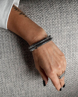 A close-up of a person's hand resting on a textured gray fabric surface, wearing a black beaded bracelet and a silver striped ring.