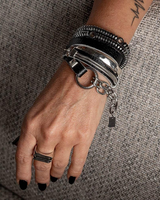 A close-up of a person's hand wearing a stack of silver-toned bracelets, including a large cuff bracelet, a coiled black bracelet, and a chain with a small padlock charm, against a textured gray fabric background.