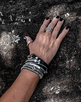 A close-up of a person's hand resting on a textured rock, showcasing a stack of bohemian-style silver and black bracelets, including a star-patterned cuff and a beaded chain bracelet.