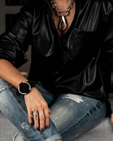 A person wearing a black leather-look button-up shirt, distressed blue jeans, a chunky silver necklace, and a black smartwatch, seated on a black-and-white checkered sofa.