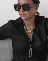 A close-up lifestyle shot of a woman wearing chunky black sunglasses with a green patterned temple, layered silver necklaces, and a silver teardrop earring, wearing a black satin shirt.