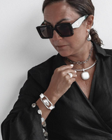 A woman wearing black sunglasses, a black satin shirt, and layered silver jewelry including a silver pendant necklace, bracelet, and earrings against a plain light gray background.
