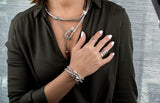 Chunky, silver-plated metal ring on an elegantly styled model's hand, paired with layered metallic necklaces and bracelets.