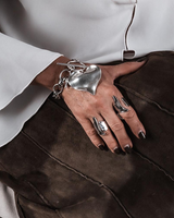 Chunky heart pendant bracelet and statement ring worn on a hand against a brown leather background, featuring a shiny metallic finish.