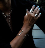 Chunky minimalist metallic cuffs featuring a wide flat adjustable cuff and an open double band cuff, worn on a hand with light blue nails.