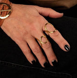 Hammered gold-plated metal ring with a geometric design, adjustable fit, worn on two fingers, with black-painted nails.