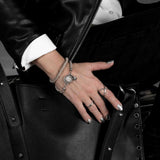 Set of silver-tone punk rings worn on a hand resting on a black leather bag, complemented by a leather jacket and silver nail polish.