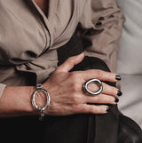 Chunky stainless steel ring with an irregular donut shape, worn on a hand with black nails, paired with a silver bracelet.
