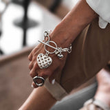 Textured heart charm bracelet with intricate woven design on an antique-plated chain, worn on a wrist alongside a chunky ring.