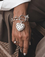 Textured heart charm bracelet in antique-plated silver, worn on a model's wrist with a toggle clasp and layered rings.