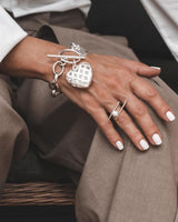 Textured heart charm bracelet with intricate details on an antique-plated chain, worn on a wrist with neutral-toned attire.