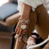 Textured heart charm bracelet with intricate heart pendant on an antique-plated chain, worn on a hand with a silver ring.