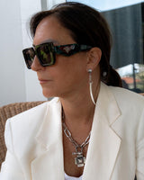 Long tiger tooth spike earrings dangle from a model's ear, showcased with a chic white blazer and bold sunglasses.