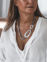 Chunky stainless steel anchor chain necklace with irregular abstract pendant, worn over a white blouse.