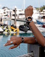 Bold wrap leather and metal cuff bracelet with sculpted silver accents, worn layered on wrist by a woman near the water.