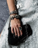 A close-up of a hand wearing a silver ring and multiple bracelets, holding a small black textured clutch bag with a silver chain strap, set against a silver sequined dress.