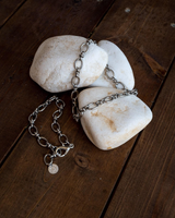 Thick stainless steel chain anklet/bracelet laid across white rocks, featuring oval links and a small charm.