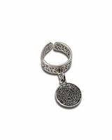 A silver-toned metal ring with an open band featuring intricate filigree cutouts, and a dangling circular charm engraved with concentric spiral patterns and text, shown on a plain white background.