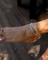 Elegant silver anklet with round charms, worn on a bare foot with white toenail polish, set against a wooden surface.