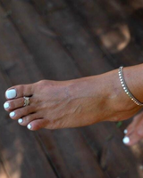 Dainty silver anklet with round charms, worn on a bare foot with white-painted toenails, against a wooden background.