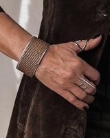 Hand wearing a textured gold bracelet and silver rings on a neutral background