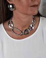 Oversized silver dome stud earrings worn with a chunky metallic necklace on a woman in a white top, showcasing a bold style.