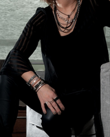 Person in a black outfit with jewelry leaning against a gray sofa in a modern room.