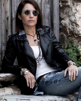 Woman wearing sunglasses and a black leather jacket sitting against a rustic wall.