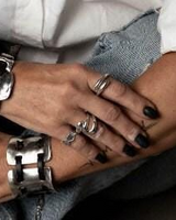 A close-up lifestyle shot of a person's hands and arms, showcasing multiple silver rings on the fingers, a chunky silver cuff bracelet on the wrist, and black nail polish, set against denim jeans and a white shirt.