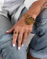 A close-up lifestyle shot of a person wearing a chunky gold beaded bracelet and matching gold rings, styled with distressed blue jeans and a white shirt.
