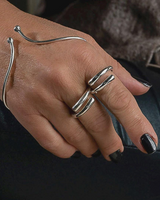 A close-up of a hand wearing minimalist silver wrap rings and a matching curved silver bracelet, with black nail polish and a dark leather surface in the background.