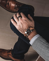 A close-up lifestyle shot of a man's hands and legs wearing a stack of beaded bracelets, a silver ring, and a gray checkered blazer, sitting on a light-colored tiled floor. He is also wearing dark trousers and brown leather dress shoes.