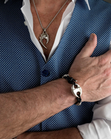 A close-up lifestyle shot of a man wearing a silver-toned geometric pendant necklace and a matching silver clasp bracelet on his wrist, styled with a blue patterned vest and white shirt.