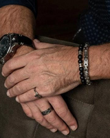 A close-up of a man's crossed arms showing several accessories, including a silver-toned watch with a black strap, a beaded bracelet, a silver chain bracelet, and rings.
