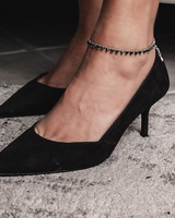 Black high-heeled shoes worn with a beaded anklet on a neutral background