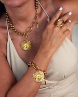 A close-up lifestyle shot of a model wearing a matching gold jewelry set, including a chunky chain necklace with a textured circular pendant, a matching bracelet, and a ring, paired with a beige halter top.