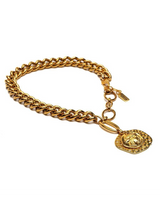A gold-tone chain bracelet with a round medallion charm featuring a raised cross, displayed on a clean white background.