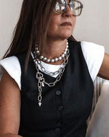 Person wearing a black vest, white shirt, and multiple necklaces with a neutral background
