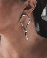 A close-up of a woman wearing a large, polished silver dangle earring with a fluid, looped organic shape.