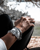 Person sitting by a body of water with hands clasped, wearing multiple bracelets.