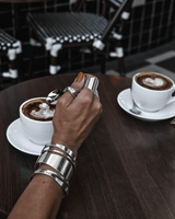 Chunky minimalist metallic cuffs set with wide flat and open double band bangles, worn on a hand holding a spoon.