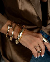 Three chunky gold cuff bangles stacked on a wrist, featuring a high-polished finish. Worn with a stylish blazer.