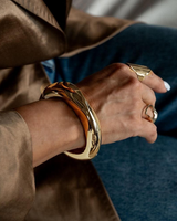 Chunky gold cuff bangle worn on wrist, featuring a polished, glossy finish, paired with additional rings.