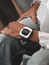 Wide black leather bracelet featuring a large silver buckle, worn on the wrist above light gray jeans.